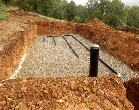 A dug trench filled with gravel and several black pipes, surrounded by earthen walls, part of a stormwater infiltration installation.
