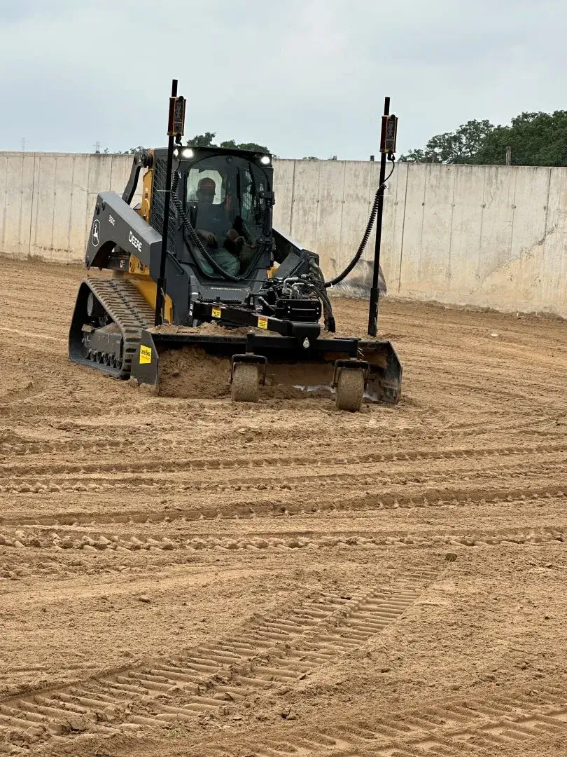 A compact track loader equipped with a GPS-guided stormwater management leveling attachment, parked on a freshly leveled dirt field.