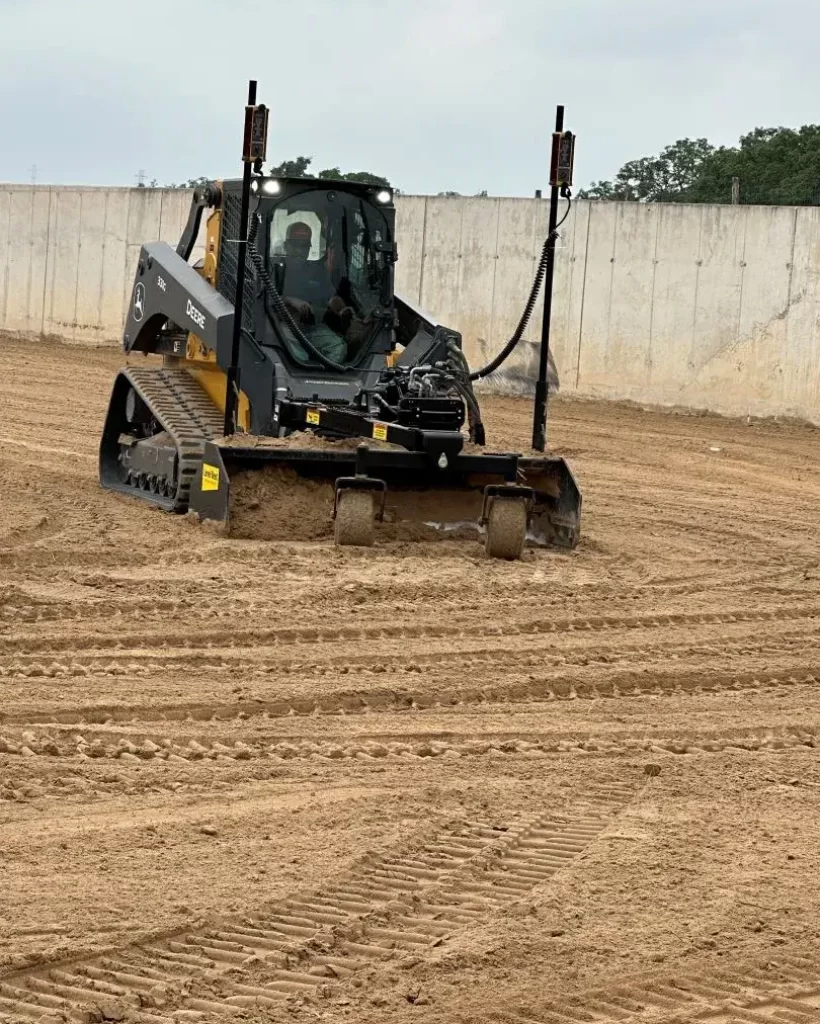 A compact track loader equipped with a GPS-guided stormwater management leveling attachment, parked on a freshly leveled dirt field.